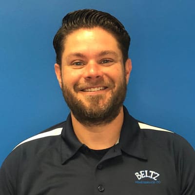 Meet The Team | Beltz Home Service Co. Man With Short Dark Hair And Beard Smiling Confidently, Embodying Team Spirit In A Navy Blue Shirt With White Accents, Positioned Against A Blue Background.