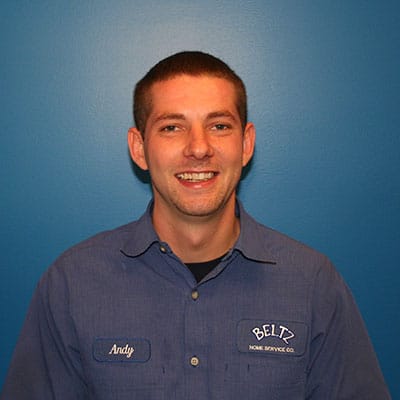 Meet The Team | Beltz Home Service Co. A Person In A Blue Uniform Shirt Stands Smiling Against A Blue Background, Embodying Team Spirit. The Shirt Features A Name Tag That Reads &Quot;Andy&Quot; Along With A Company Logo, Reflecting An Inclusive Work Environment.