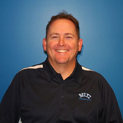 Meet The Team | Beltz Home Service Co. A Smiling Team Member Wearing A Black Polo Shirt With A White Logo Stands Confidently Against A Soothing Blue Background.