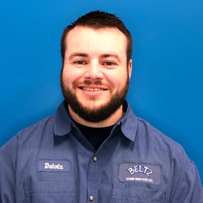 Meet The Team | Beltz Home Service Co. A Person With A Beard And Short Hair Is Smiling, Wearing A Blue Shirt Adorned With Company Logos And A Name Tag, Embodying The Spirit Of The Team. The Background Is Solid Blue.