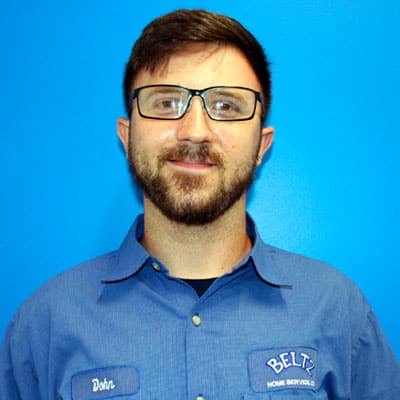 Meet The Team | Beltz Home Service Co. A Person With Glasses And A Beard, Wearing A Blue Button-Up Shirt, Stands Confidently Against A Blue Background, Embodying The Spirit Of Teamwork.