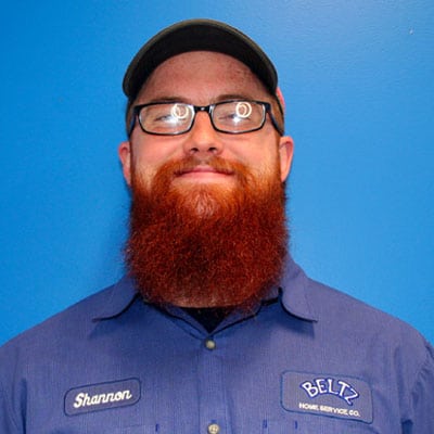 Meet The Team | Beltz Home Service Co. A Person With Glasses And A Reddish Beard, Wearing A Blue Shirt And Cap, Stands Against A Bright Blue Background. The Shirt Features The Name &Quot;Shannon&Quot; And A &Quot;Beltz Home Service Co.&Quot; Logo, Proudly Representing The Team'S Dedication To Quality Service.