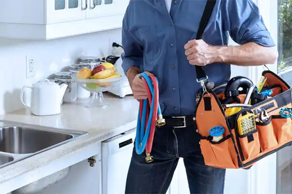 A service technician is in a residential kitchen with a bag of tools near a sink.