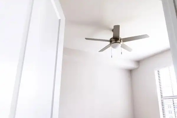 An all white room with a window and alone ceiling fan.