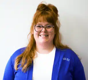 Meet The Team | Beltz Home Service Co. A Person With Long Red Hair And Glasses, Smiling Warmly, Stands Proudly Against A Plain White Background, Wearing A Blue Sweater Over A White Shirt—A True Team Player Showcasing Their Approachable And Friendly Demeanor.