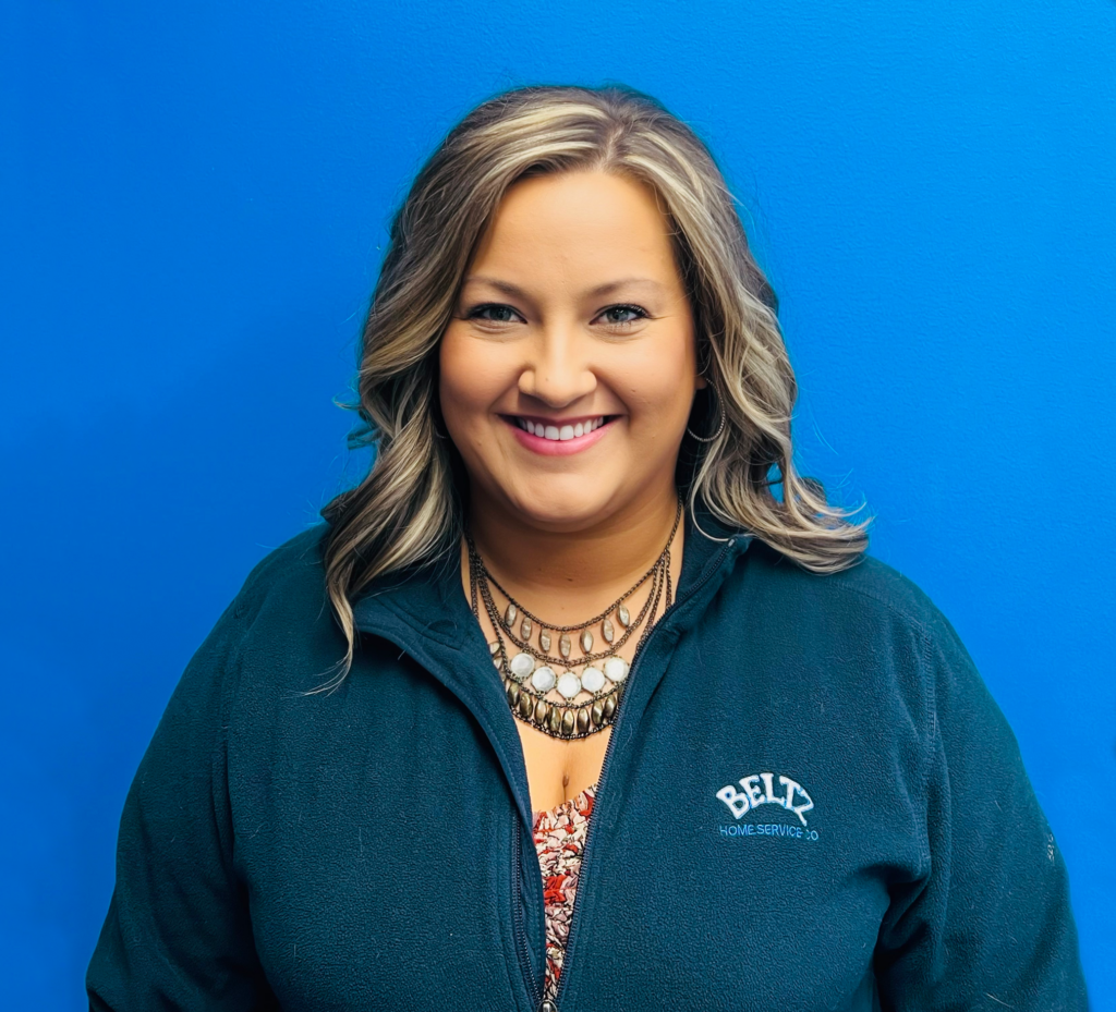 Meet The Team | Beltz Home Service Co. A Person With Long Hair And A Necklace Smiles Against A Bright Blue Background, Proudly Wearing A Dark Green Fleece Jacket Embroidered With &Quot;Belt Home Service Co.,&Quot; Showcasing Their Dedication As Part Of The Team.