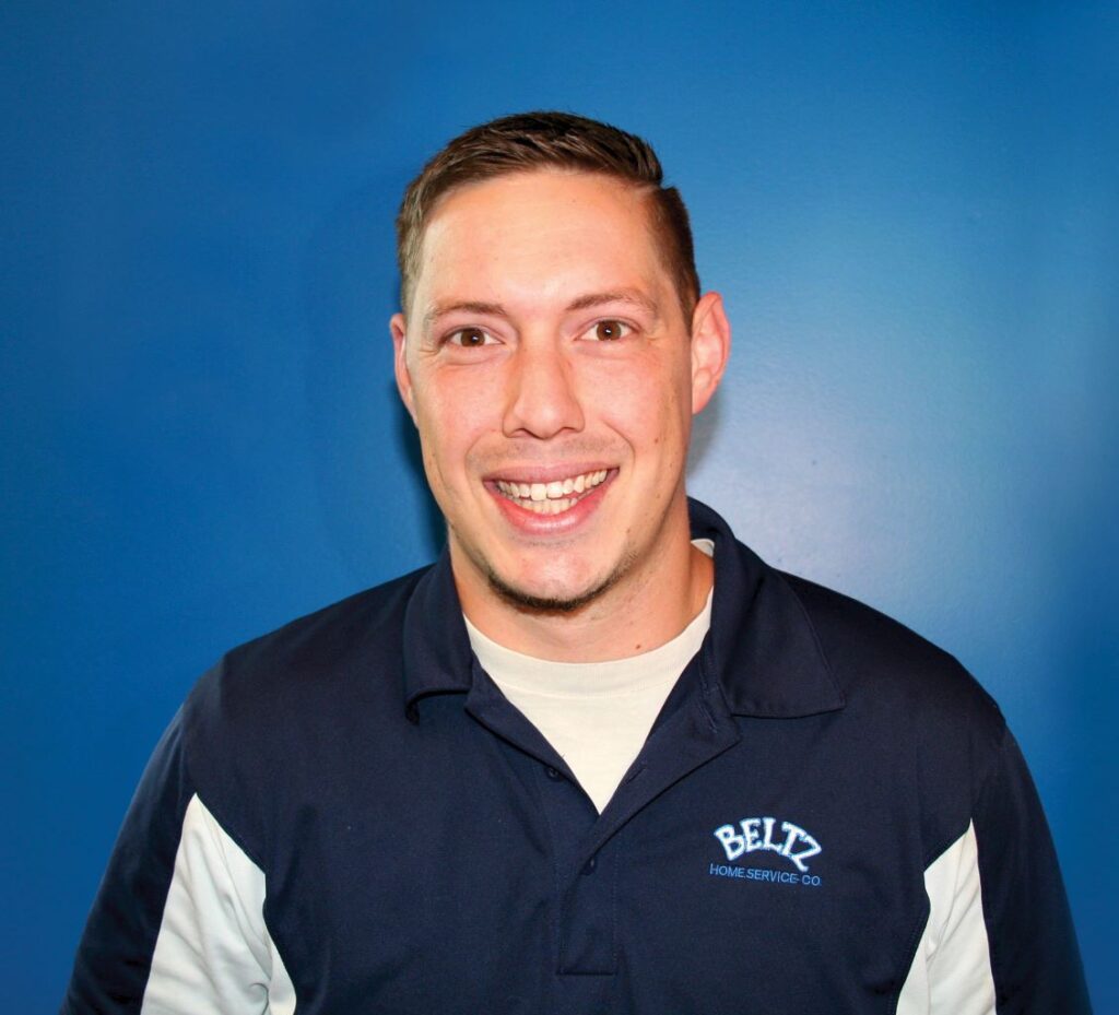 Meet The Team | Beltz Home Service Co. A Person Smiling In A Navy Shirt With White Details Stands Confidently In Front Of A Solid Blue Background, Exuding A Sense Of Team Spirit.