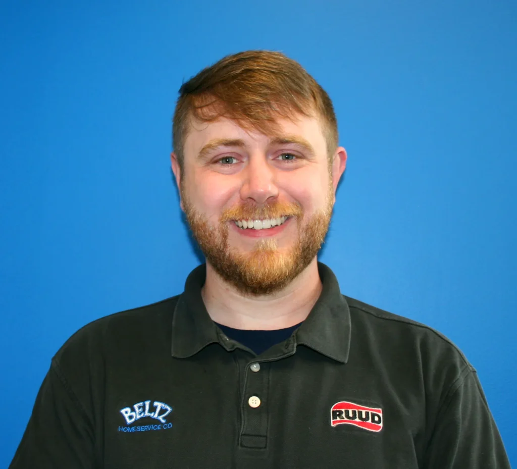 Meet The Team | Beltz Home Service Co. A Smiling Man With Short Brown Hair And A Beard, Wearing A Black Polo Shirt Featuring &Quot;Beltz Home Service Co&Quot; And &Quot;Ruud&Quot; Logos, Stands Proudly As Part Of The Team In Front Of A Blue Wall.