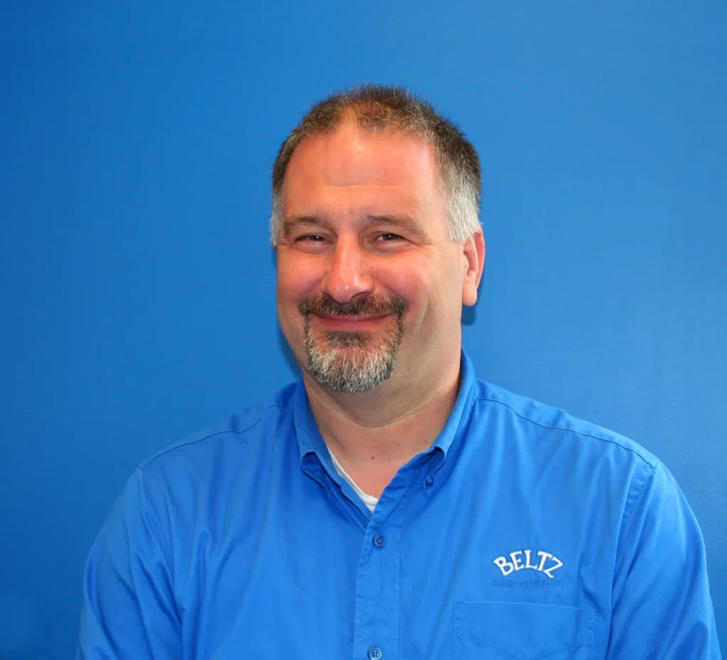 Meet The Team | Beltz Home Service Co. A Man With Short Hair And A Goatee, Wearing A Blue &Quot;Beltz&Quot; Team Shirt, Smiles In Front Of A Solid Blue Background.