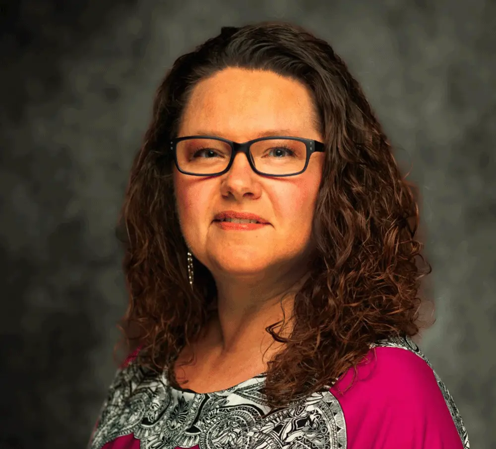 Meet The Team | Beltz Home Service Co. A Woman With Curly Brown Hair And Glasses, Wearing A Pink And Patterned Blouse, Poses Confidently In Front Of A Dark Mottled Background, Ready To Join The Team.