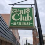 Green And White Sign For “The 818 Club Restaurant” Hangs From A Building On A Cloudy Day, With Nearby Buildings And A Church Visible In The Background.