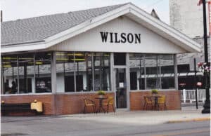 A Restaurant With Large Windows, Labeled “Wilson” On The Front. Two Small Tables With Chairs And Potted Plants Are Set On The Sidewalk Outside The Entrance.