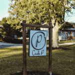 A Wooden Sign In Grassy Area Reads “Circle P” With A Large Letter “P” Inside A Circle, Trees And A Building In The Background.