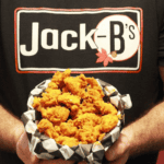 Person Holding A Basket Of Fried Chicken Pieces In Front Of Their Chest, Wearing A Black T-Shirt With “Jack-B’s” Printed On It.