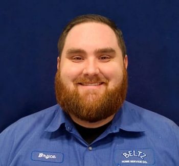 Bryce A Bearded Person Smiles Warmly Against A Dark Blue Background, Showcasing A Blue Shirt Adorned With A Name Tag And The Company Logo, Embodying The Spirit Of Teamwork.