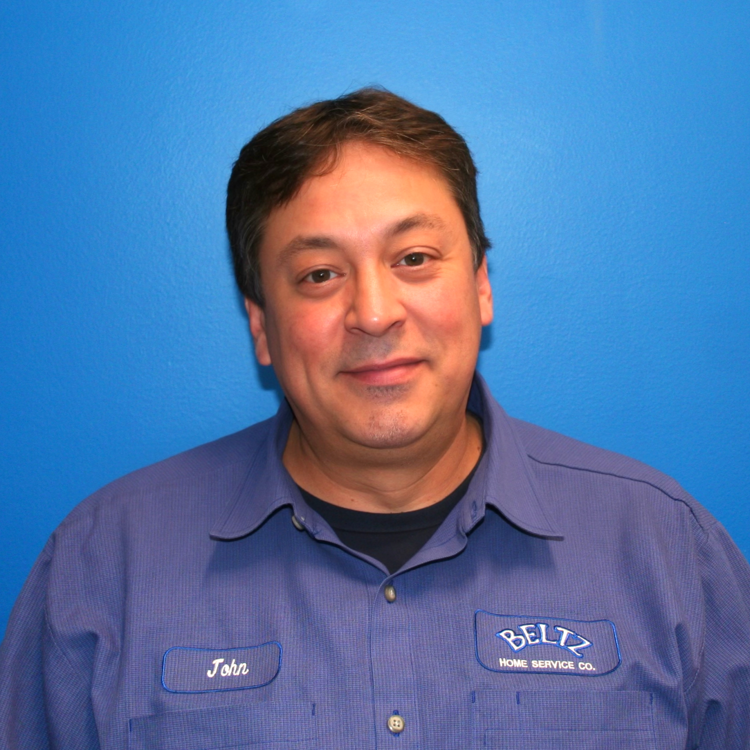 John A Team Member In A Blue Work Shirt With A Name Tag Stands Against A Bright Blue Background.