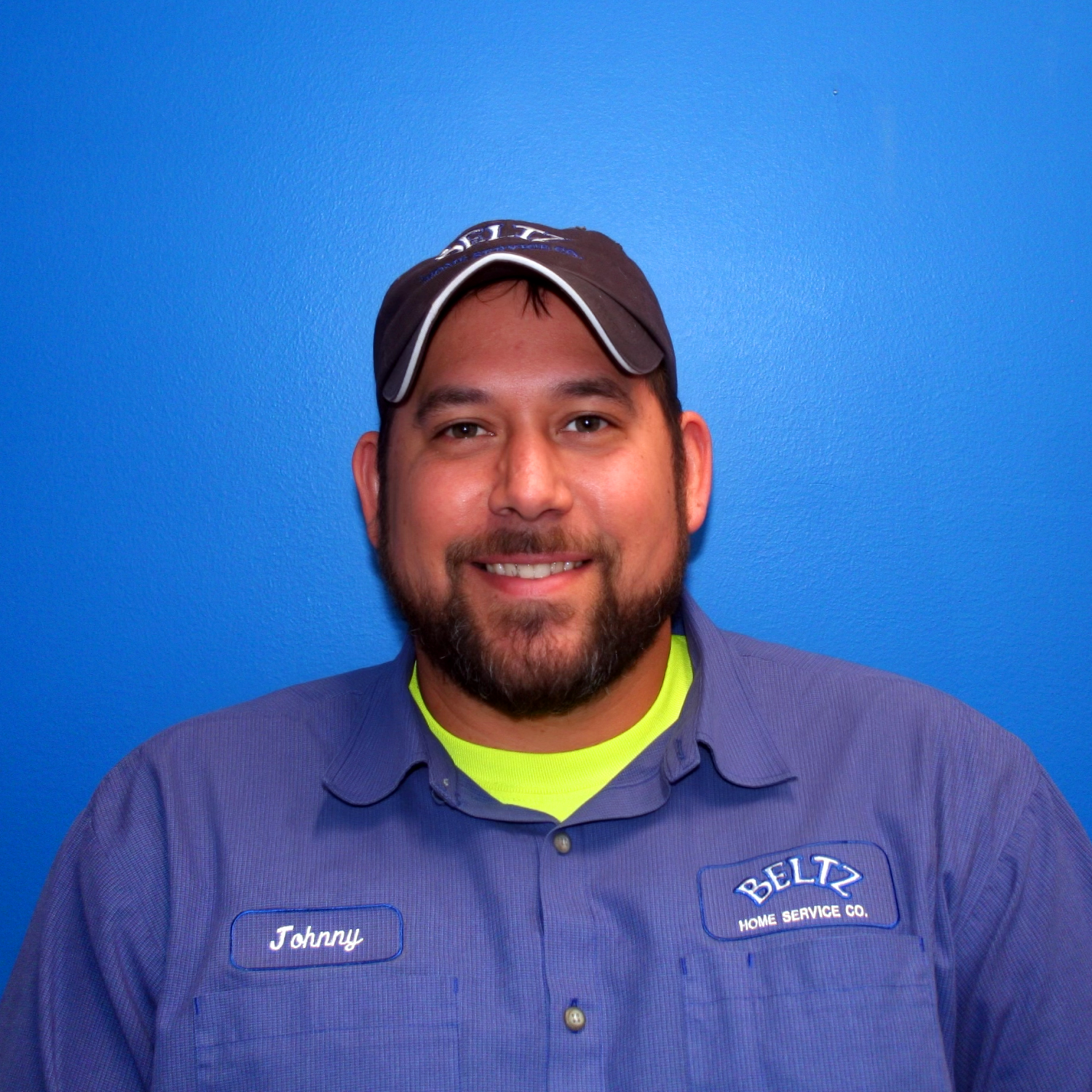 Johnny A Man With A Beard Is Smiling, Wearing A Cap And A Blue Work Shirt With The &Quot;Beltz Home Service Co.&Quot; Logo And A Name Patch That Reads &Quot;Johnny.&Quot; He Represents The Dedicated Team Spirit Against A Bright Blue Background.