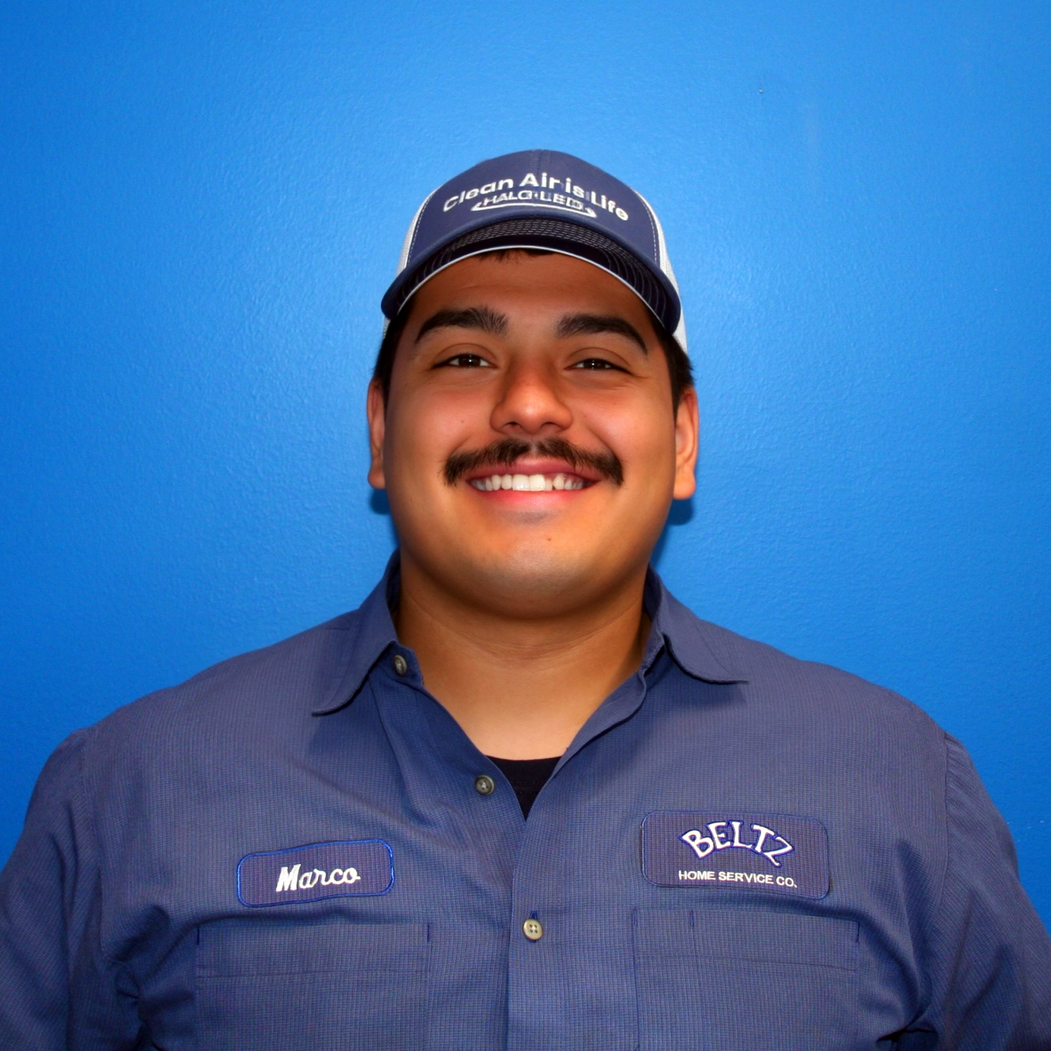 Marco A Person Smiling In A Blue Uniform And Cap, Standing Against A Bright Blue Background. The Uniform'S Name Patches Read &Quot;Marco&Quot; And &Quot;Beltz Home Service Co.,&Quot; Proudly Representing The Dedicated Team Behind Every Service.