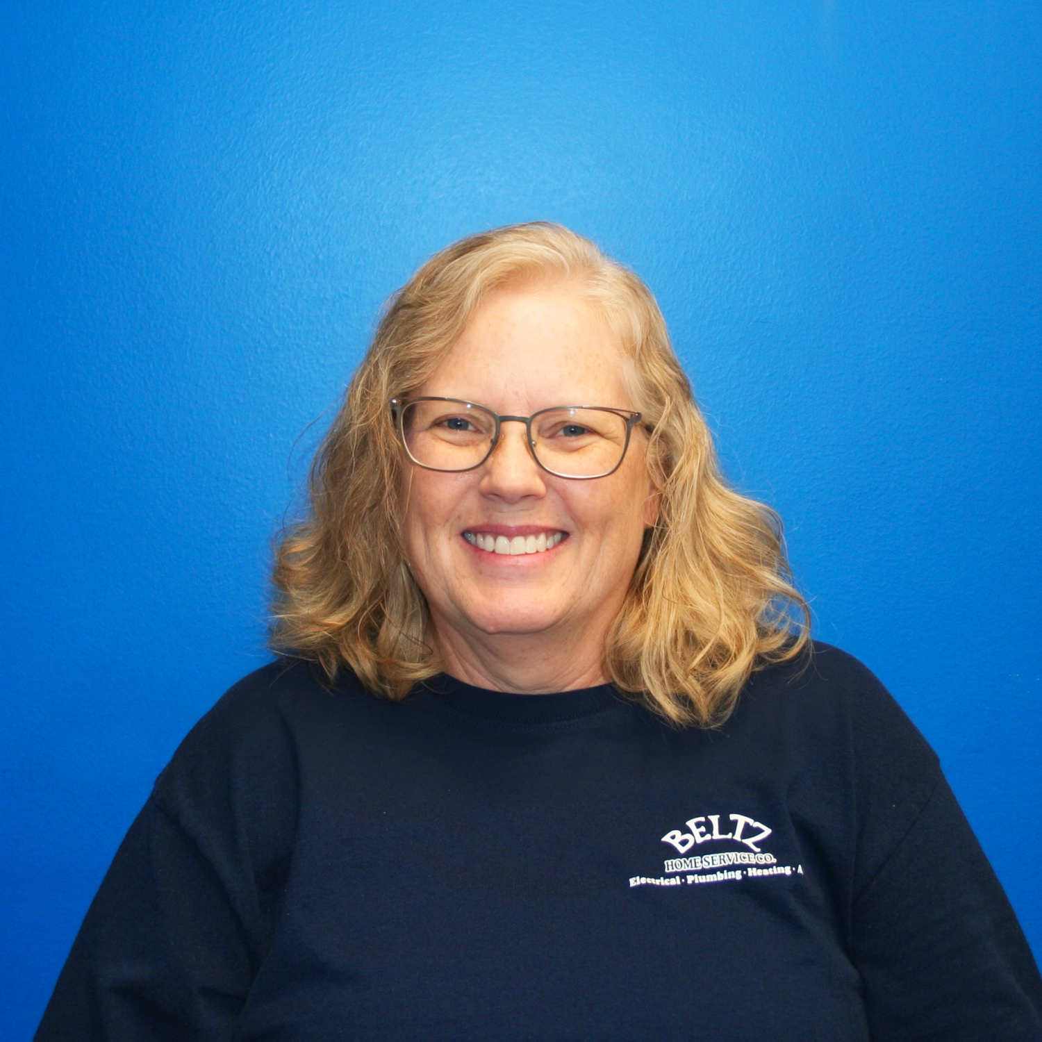 Teresa A Smiling Person With Glasses And Shoulder-Length Blond Hair, Wearing A Navy T-Shirt Featuring A Team Logo, Stands Against A Blue Background.